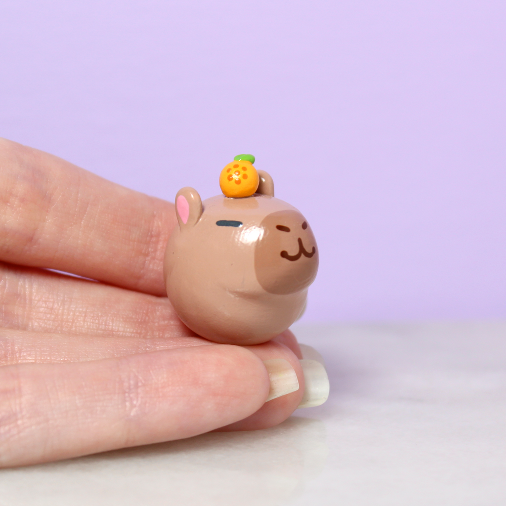 A hand holding a capybara figurine. The capybara is one inch tall and has a tiny orange on its head.