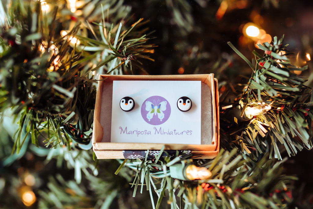 A pair of penguin stud earrings nestled in a jewelry box and sitting in the branches of a christmas tree.