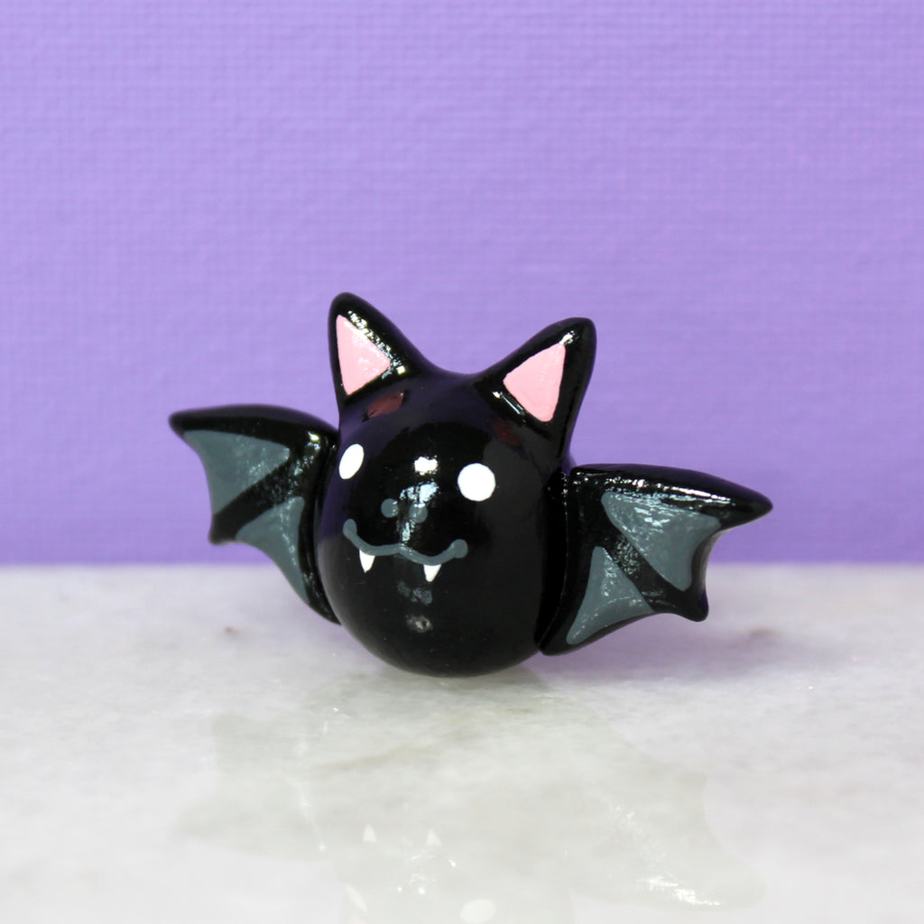 A small black bat figurine sitting on a shiny marble surface with a purple background. The figurine is glossy and shiny, and has a sweet toothy smile.