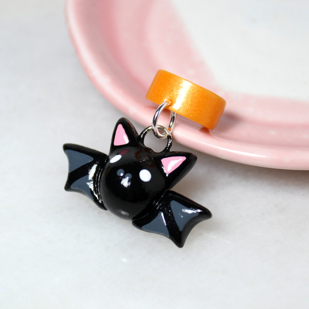 A black bat charm hangs from a sparkly orange ear cuff. The ear cuff rests against a pink and white jewelry dish.