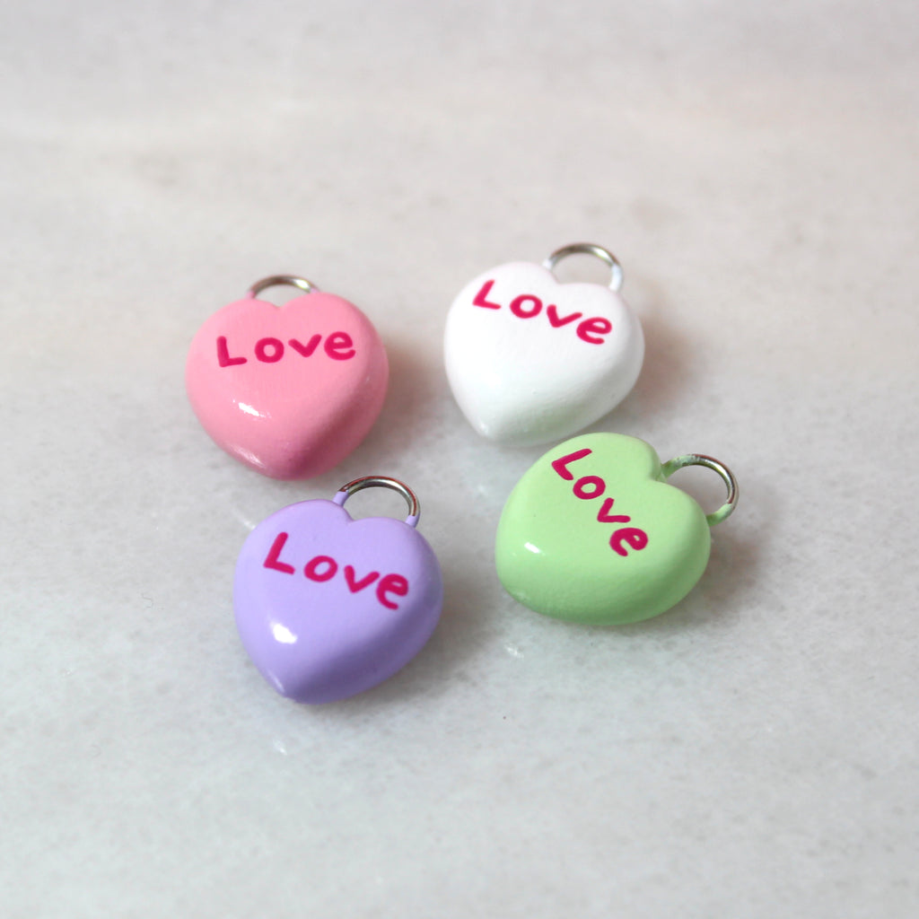 Four pastel candy heart charms. Each charm has the text "Love" written on them. The charms come in white, pink, green, and purple.