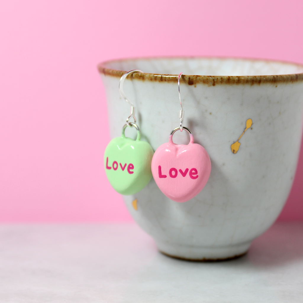 A pair of mismatched candy heart earrings hang from a white cup. The charms are attached to sterling silver ear wires.