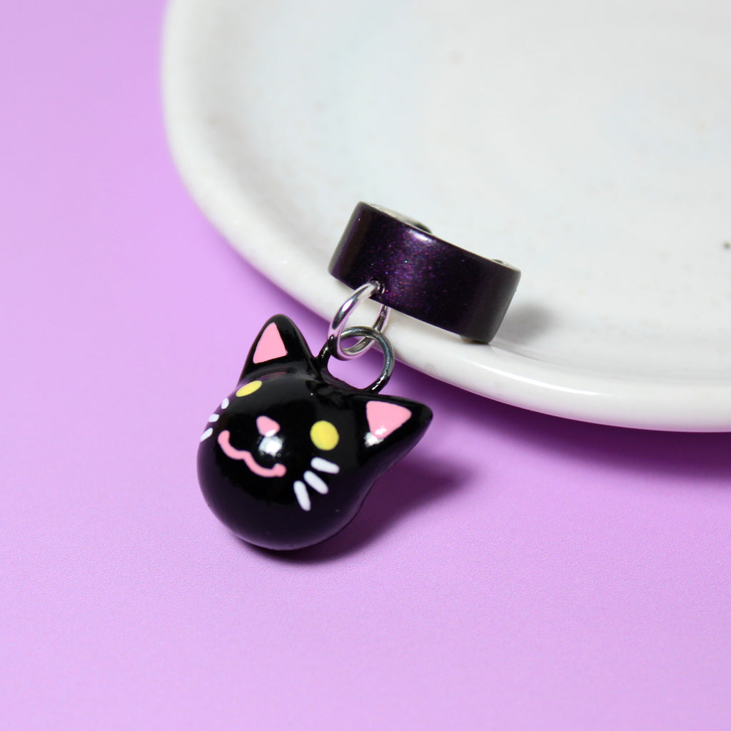 A black cat charm hangs from a sparkly dark purple ear cuff. The ear cuff rests against a white jewelry dish.