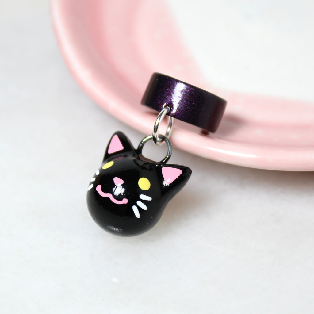 A black cat charm hangs from a sparkly dark purple ear cuff. The ear cuff rests against a white jewelry dish.