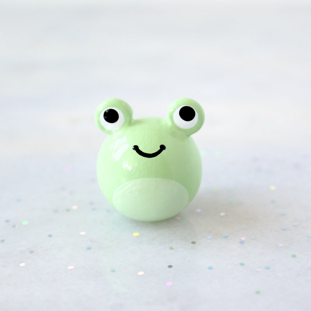 A chubby green frog figurine with big eyes and a wide happy smile. The figurine is sitting on a marble surface with rainbow glitter.