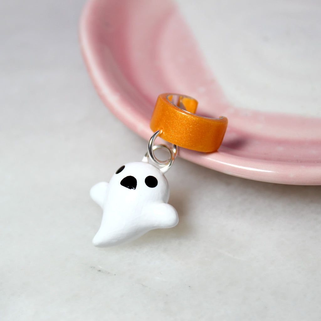A ghost charm hangs from a sparkly orange ear cuff. The ear cuff rests against a pink jewelry dish.