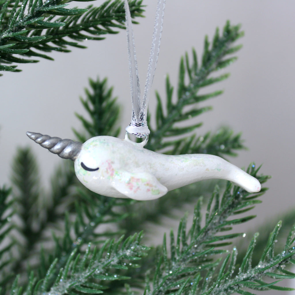 A white narwhal ornament with iridescent flakes hangs on a christmas tree.