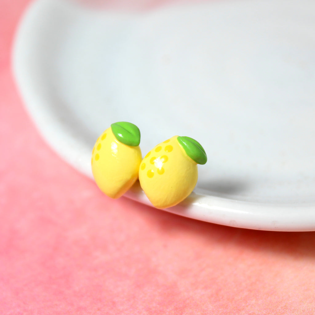 A pair of tiny yellow lemon earrings rest on the rim of a white dish. The dish is on top of a watercolor pink background.