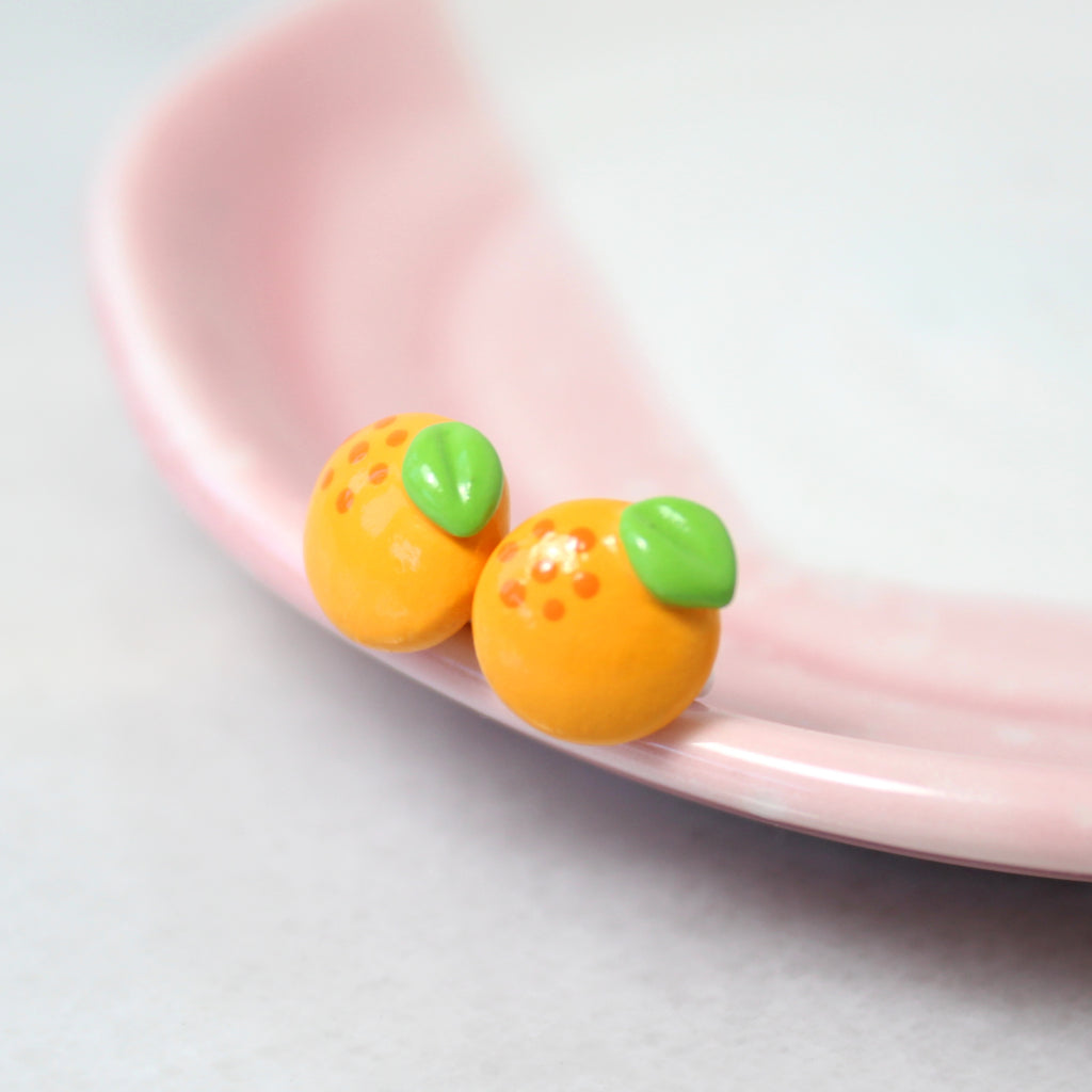 A pair of orange fruit earrings rest on the rim of a pink and white dish.