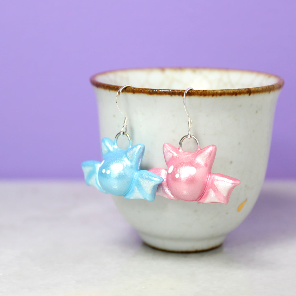 A pair of pastel bat earrings hang from a white cup. The bat on the left is sparkly baby blue and the bat on the right is sparkly bubblegum pink.