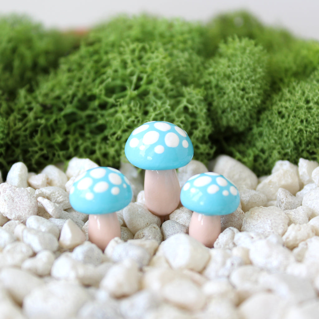 Three turquoise blue mushrooms with white spots are sticking out of a planter pot filled with pebbles and moss.