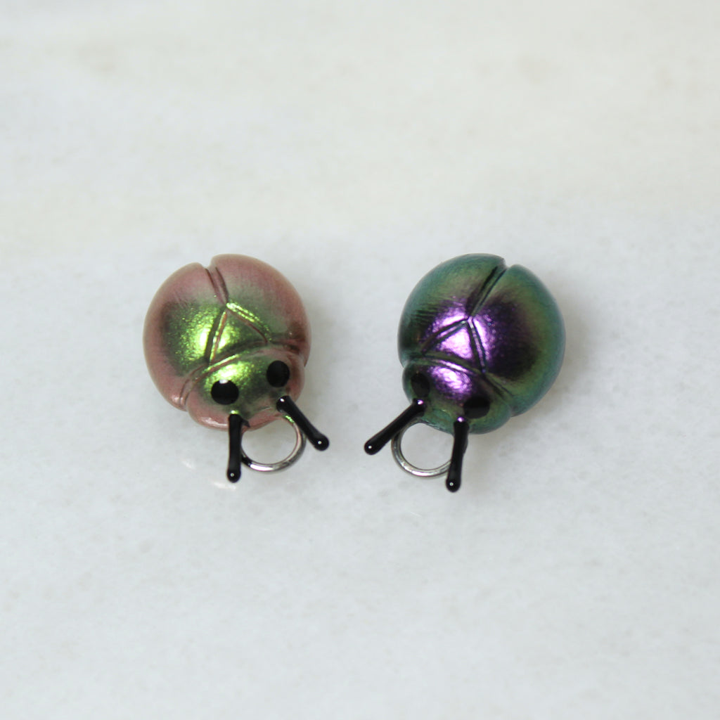Two iridescent beetle charms sit on a marble surface. One beetle is a sparkly green/gold, and the other is a sparkly purple/silver