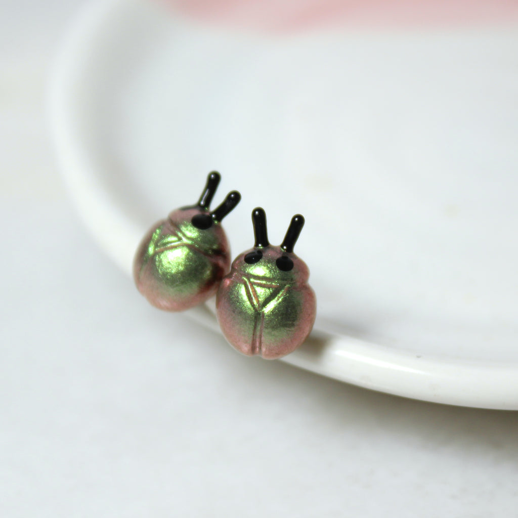 A pair of iridescent green and gold beetle earrings rest against a white jewelry dish