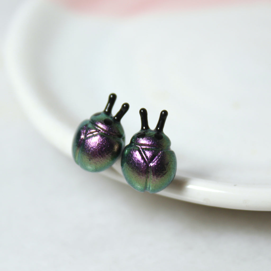 A pair of iridescent purple and silver beetle earrings rest against a white jewelry dish