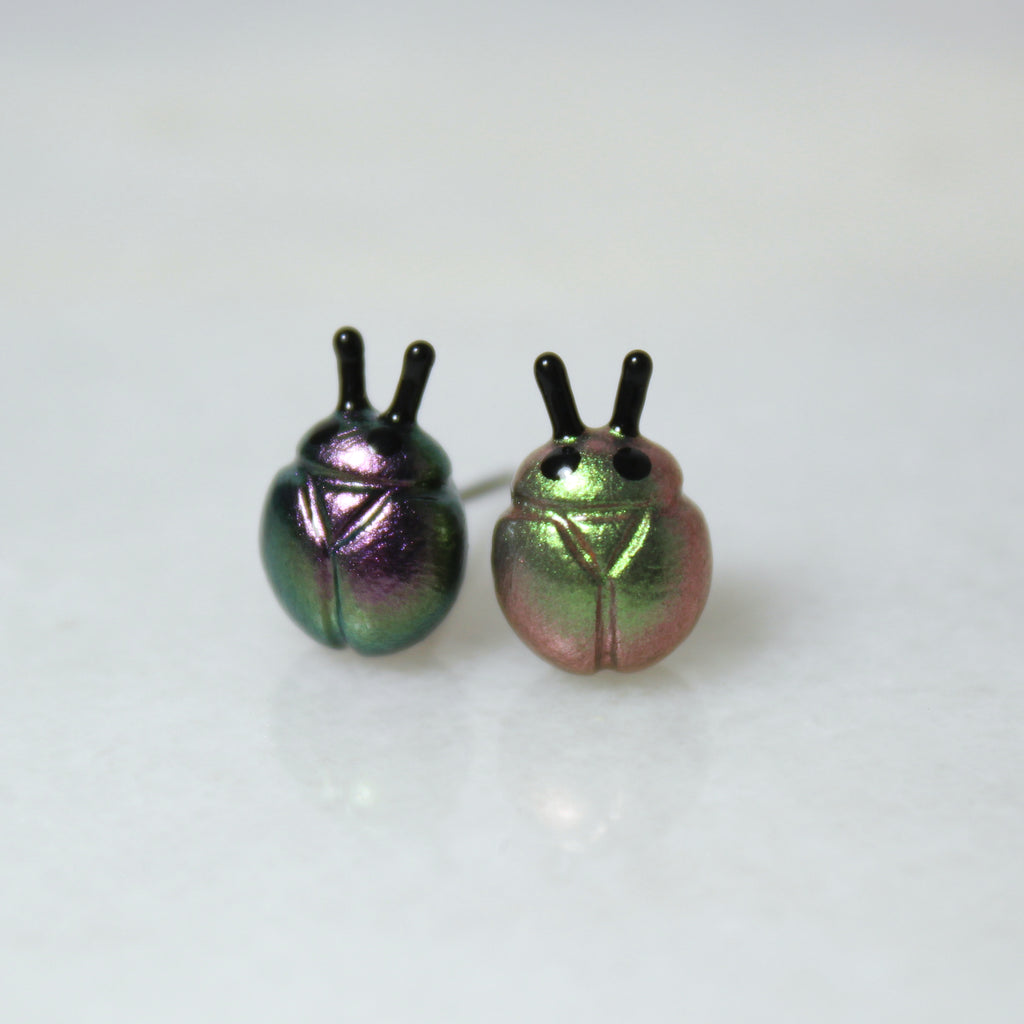 A purple and silver beetle earring sits next to a green and gold beetle earring