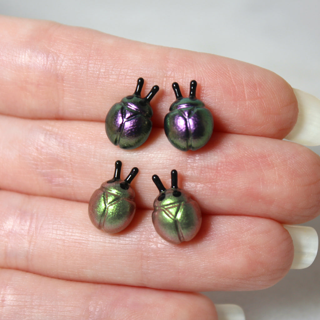 A hand holds two pairs of iridescent beetle earrings. The top earrings are purple and silver while the bottom earrings are green and gold