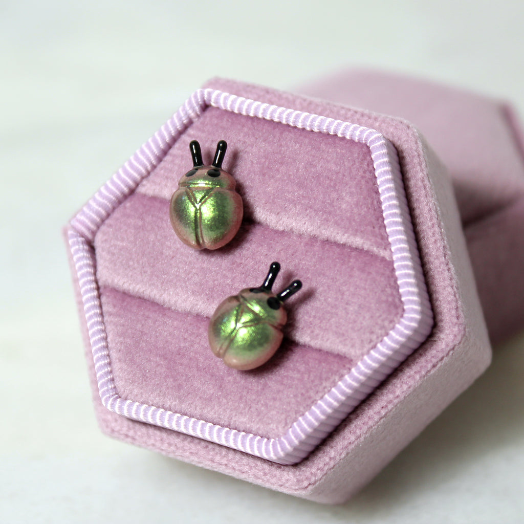 A pair of iridescent green and gold beetle earrings are displayed in a purple jewelry box