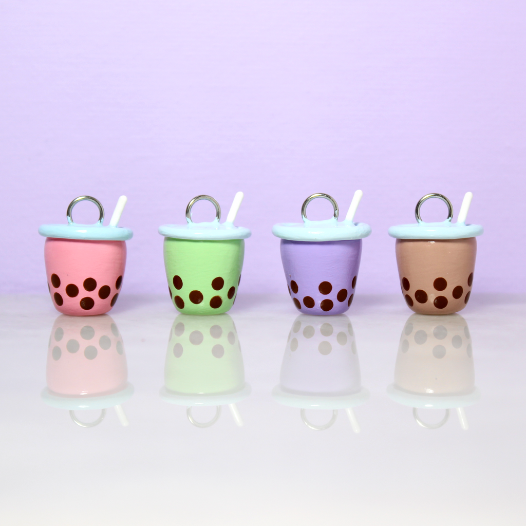 Four bubble tea charms are lined up in a row. From left to right is a pink charm, a green charm, a purple charm, and a brown charm.