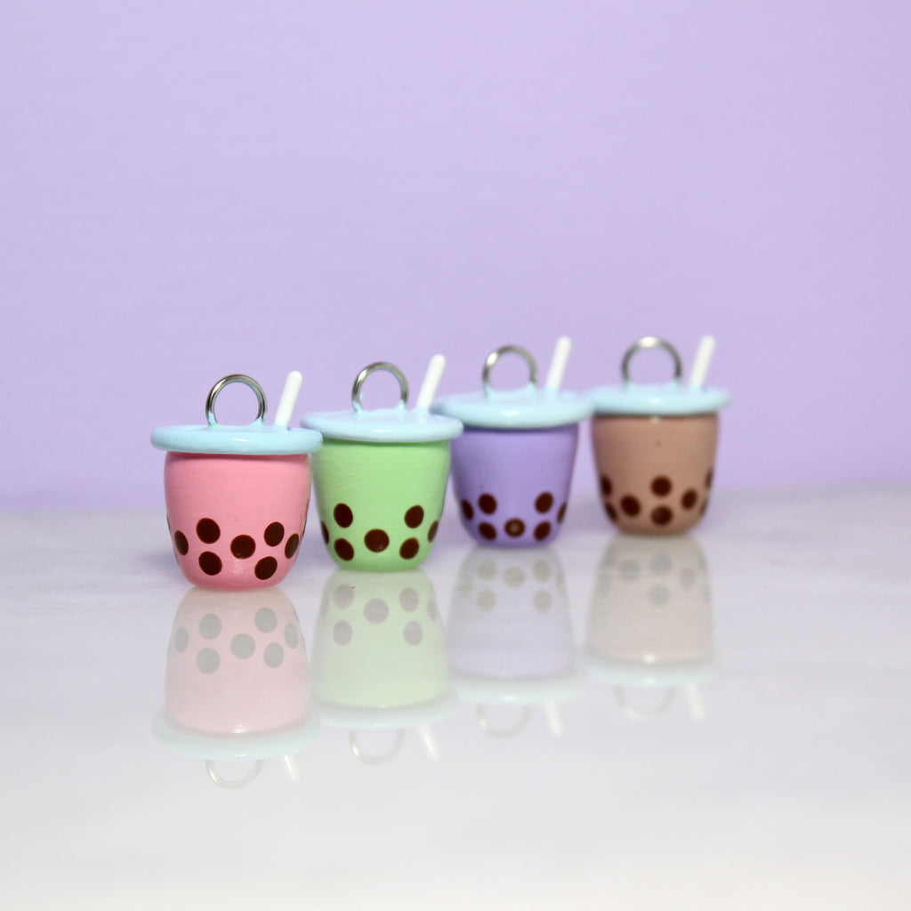 Four bubble tea charms are lined up in a row. From left to right is a pink charm, a green charm, a purple charm, and a brown charm.