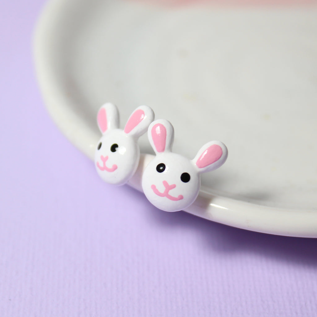 A pair of white bunny stud earrings rest against a white jewelry dish