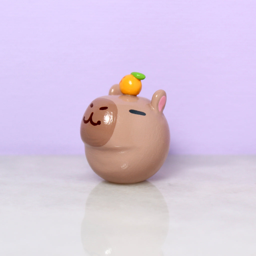 A chubby capybara figurine with a tiny orange on its head.