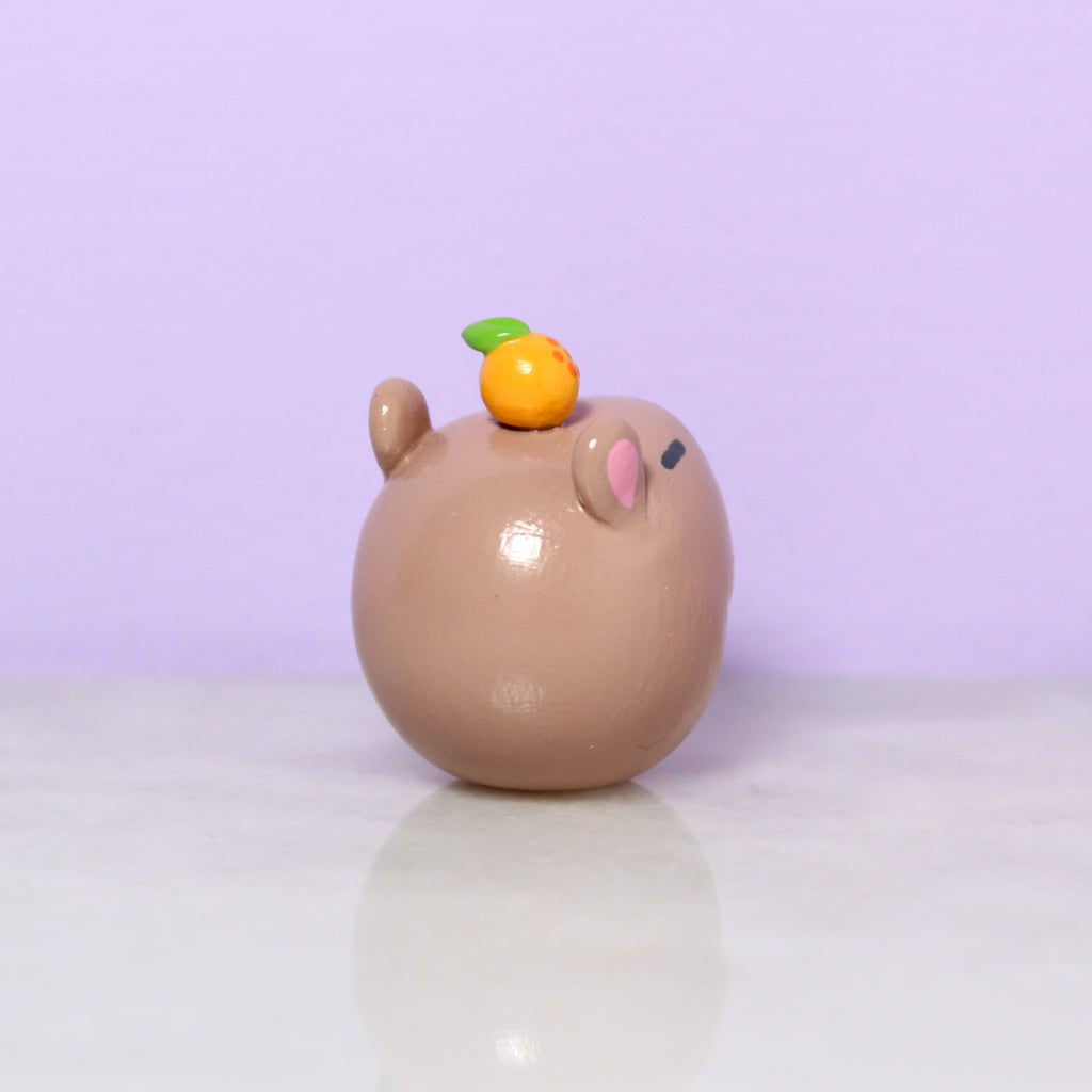 Backside of a chubby capybara figurine with a tiny orange on its head.