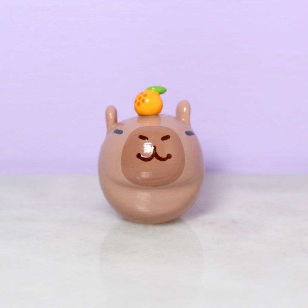 A chubby capybara figurine with a tiny orange on its head.