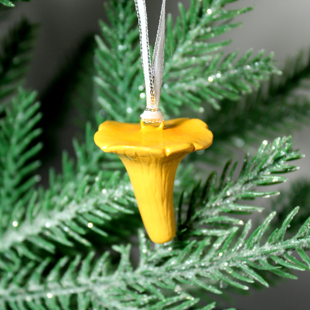 A yellow and orange chanterelle mushroom ornament hangs from a silver ribbon on a pine tree