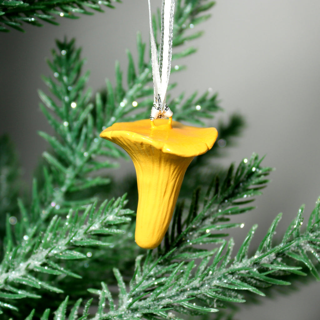 A yellow and orange chanterelle mushroom ornament hangs from a silver ribbon on a pine tree