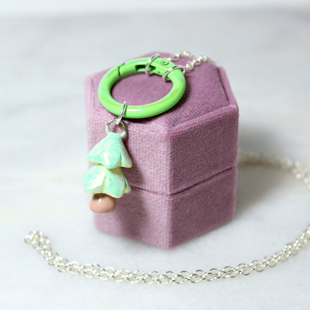 A pastel green christmas tree charm hangs from a matching green carabiner necklace. The necklace hangs off of a purple jewelry box