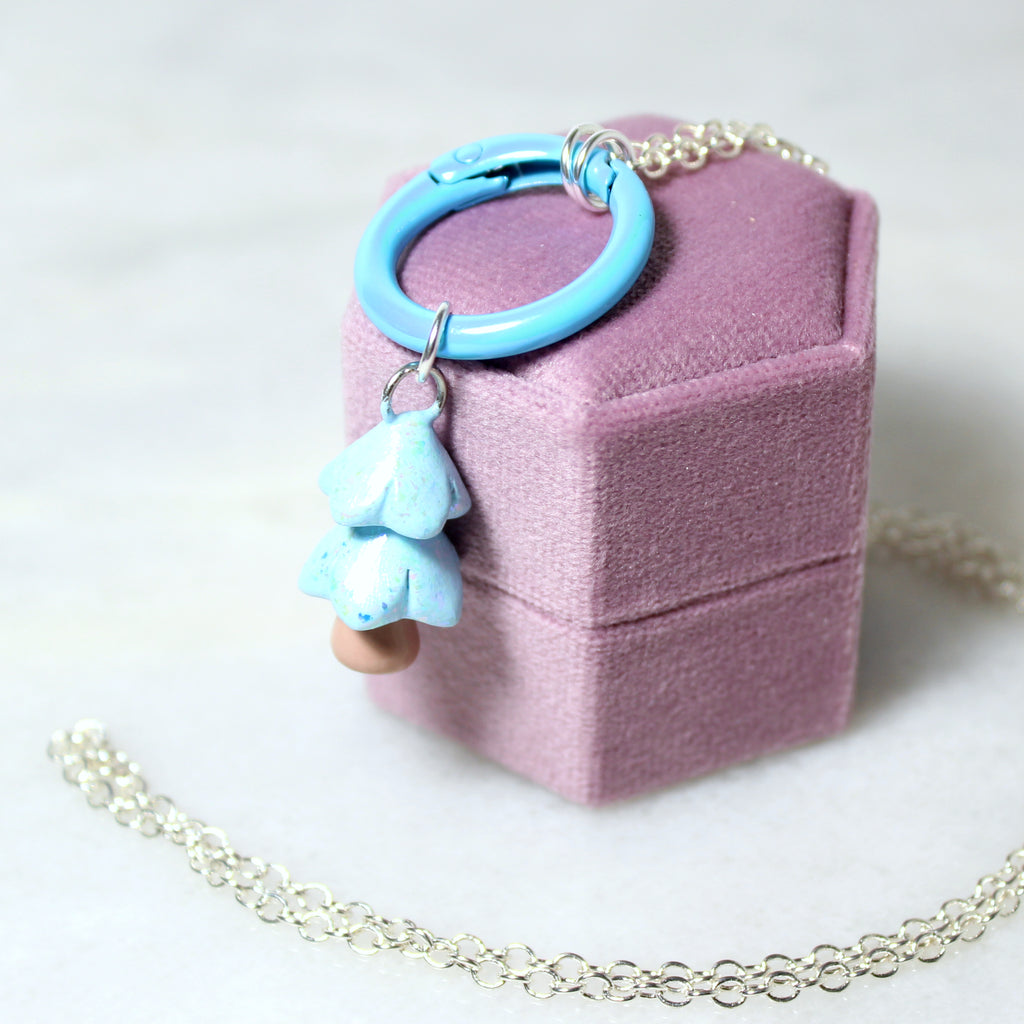 A pastel blue christmas tree charm hangs from a matching blue carabiner necklace. The necklace hangs off of a purple jewelry box