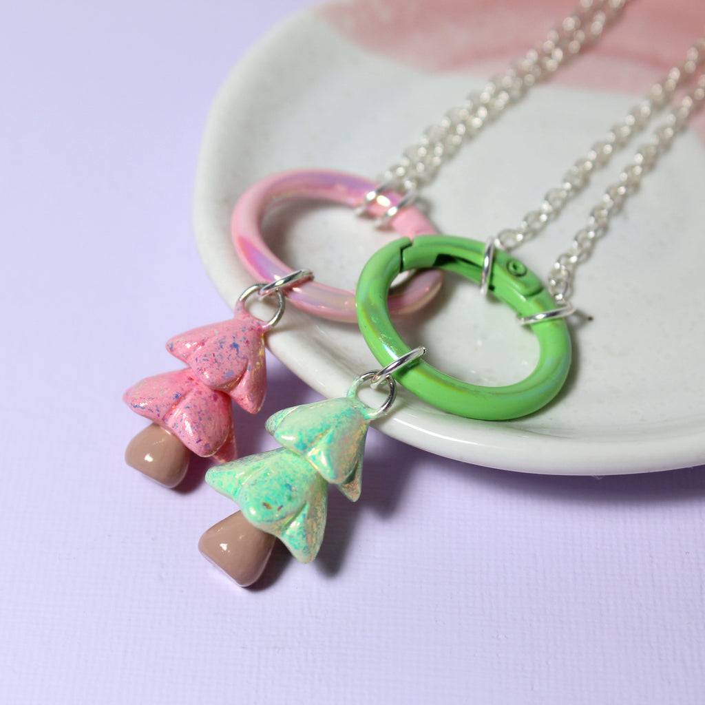 Two sparkly pastel christmas tree charms with matching carabiner charm keeper clasps rest against a white jewelry dish
