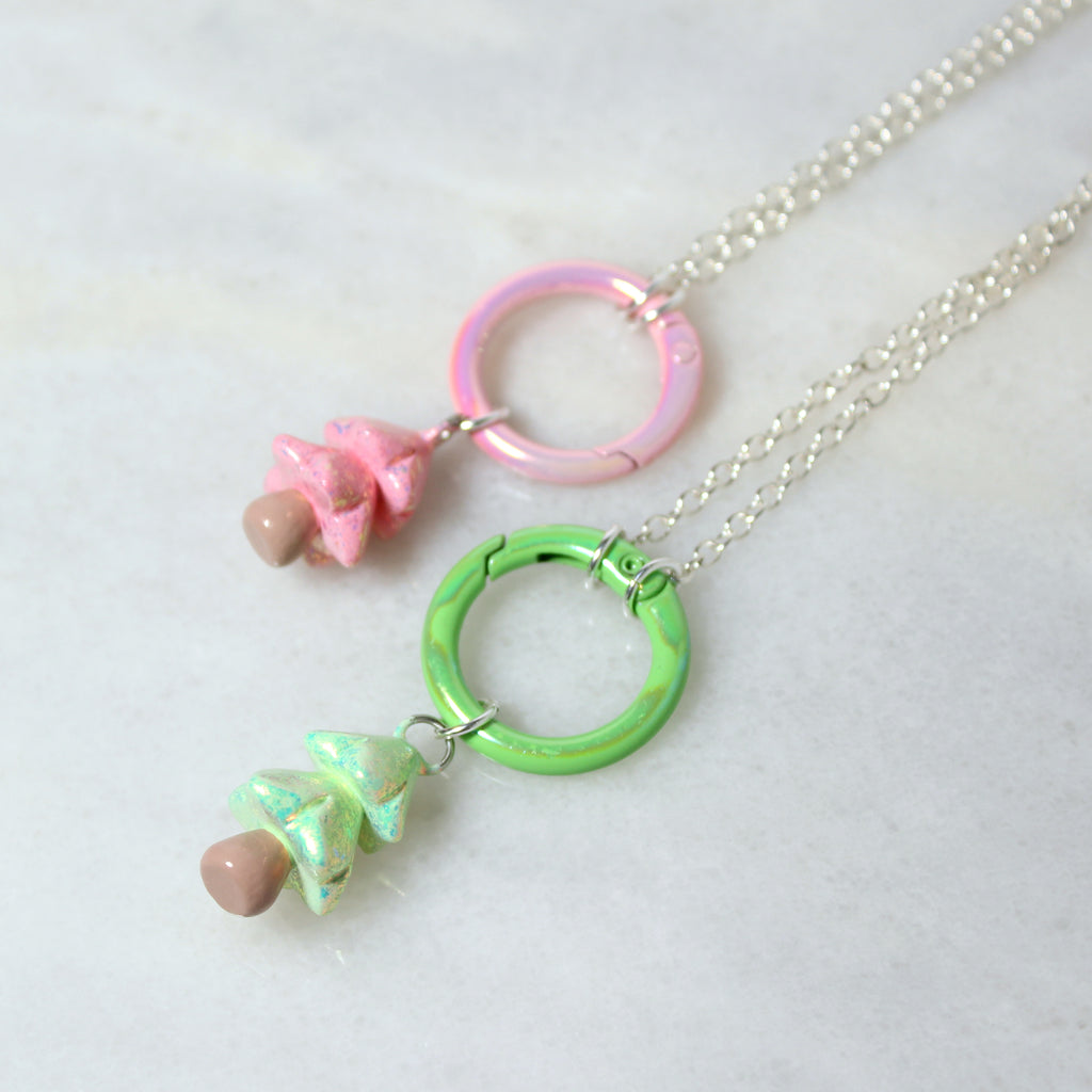 A sparkly pink and a sparkly green christmas tree necklace with large pastel carabiner clasps rest against a marble surface