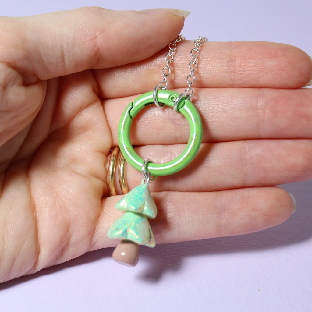A hand holds a sparkly christmas tree charm necklace with a large round carabiner charm keeper clasp