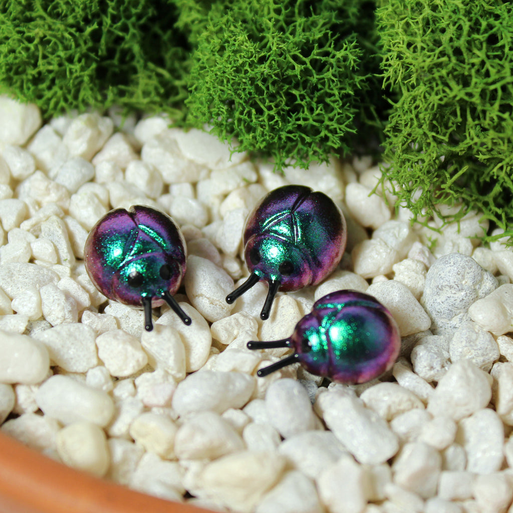 Three miniature beetle figurines sit in a moss garden. The beetles have iridescent teal/purple shells