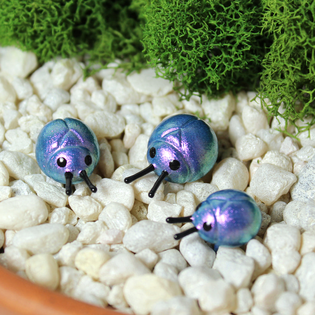 Three miniature beetle figurines sit in a moss garden. The beetles have iridescent lavender/light blue shells