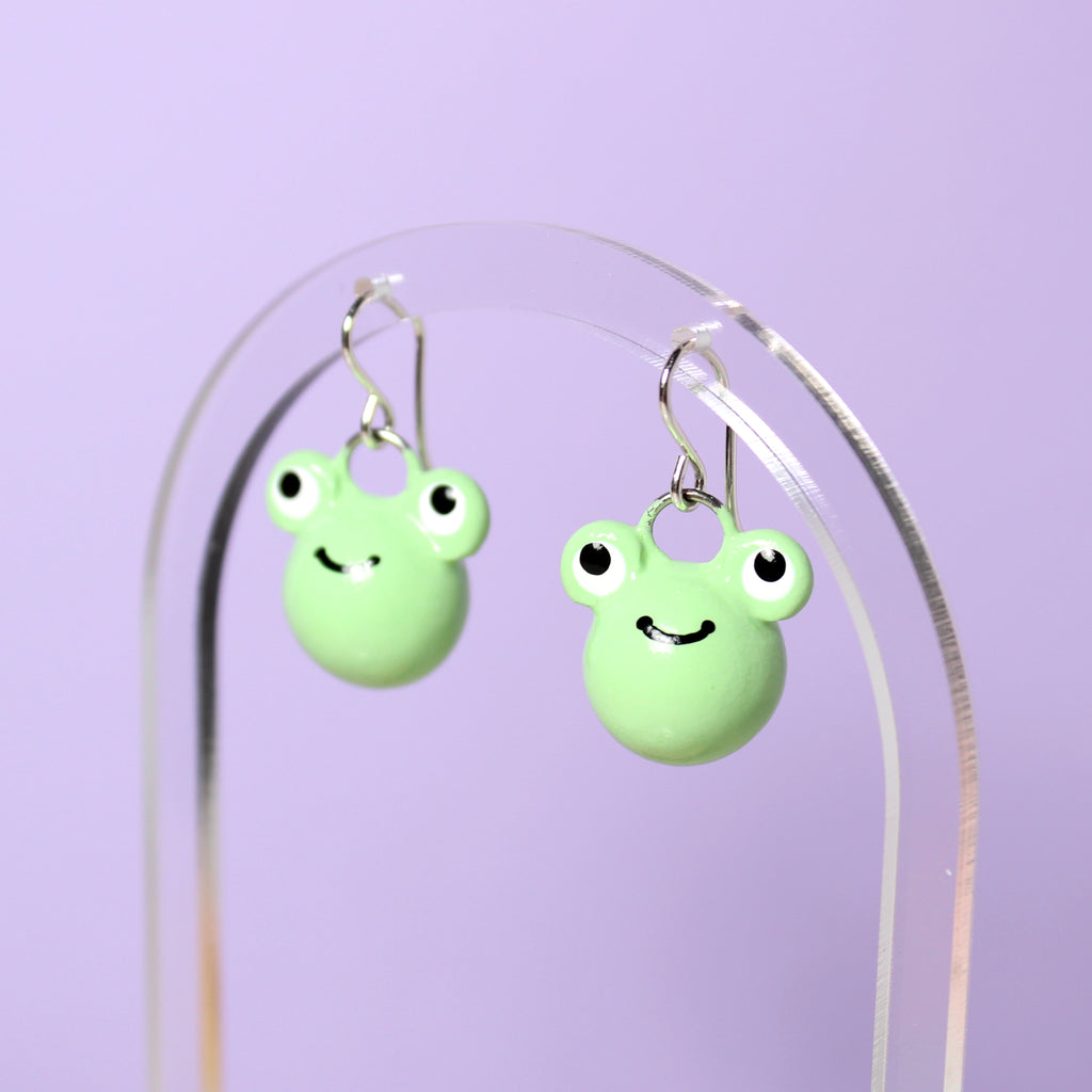 A pair of frog earrings hang from a clear acrylic earring display