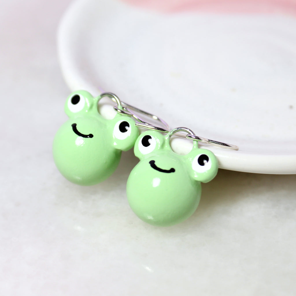 A pair of smiling green frog charms hang from sterling silver earring hooks. The earrings rest against a white jewelry dish