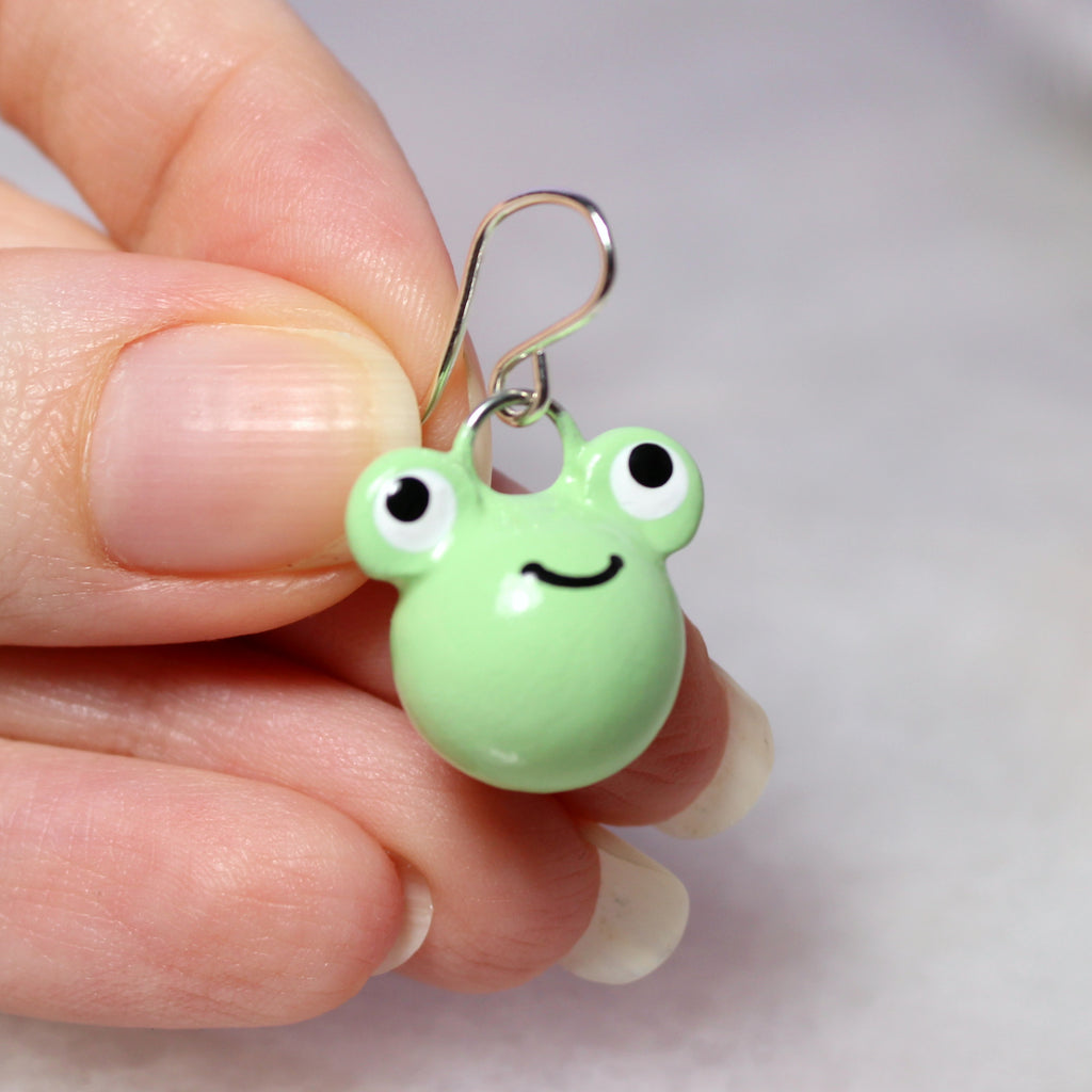 A hand holds a single frog earring by the sterling silver earring hook. The frog charm is about 3/4 of an inch tall