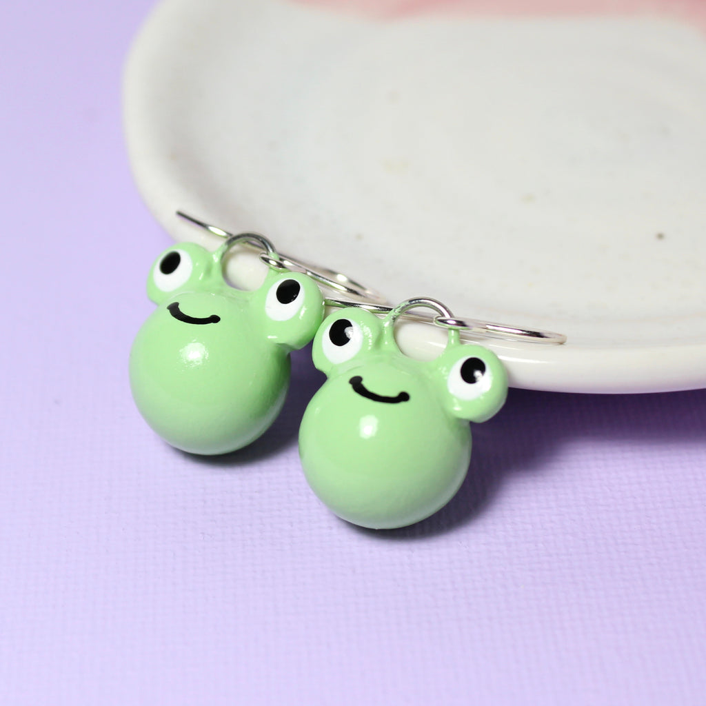 A pair of smiling green frog charms hang from sterling silver earring hooks. The earrings rest against a white jewelry dish