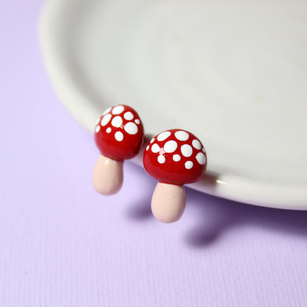 A pair of red mushroom earrings with white speckles rest against a white jewelry dish