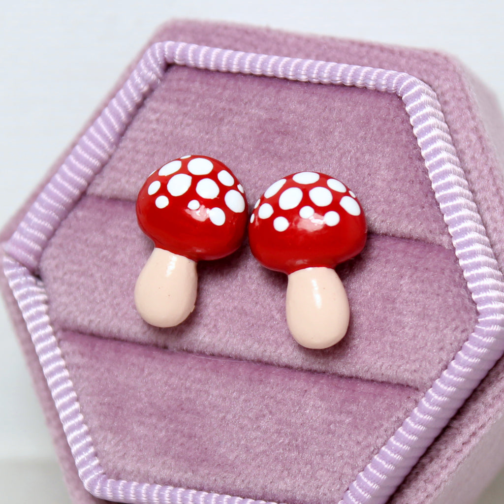 A pair of red mushroom stud earrings are nestled in a hexagonal purple velvet jewelry box
