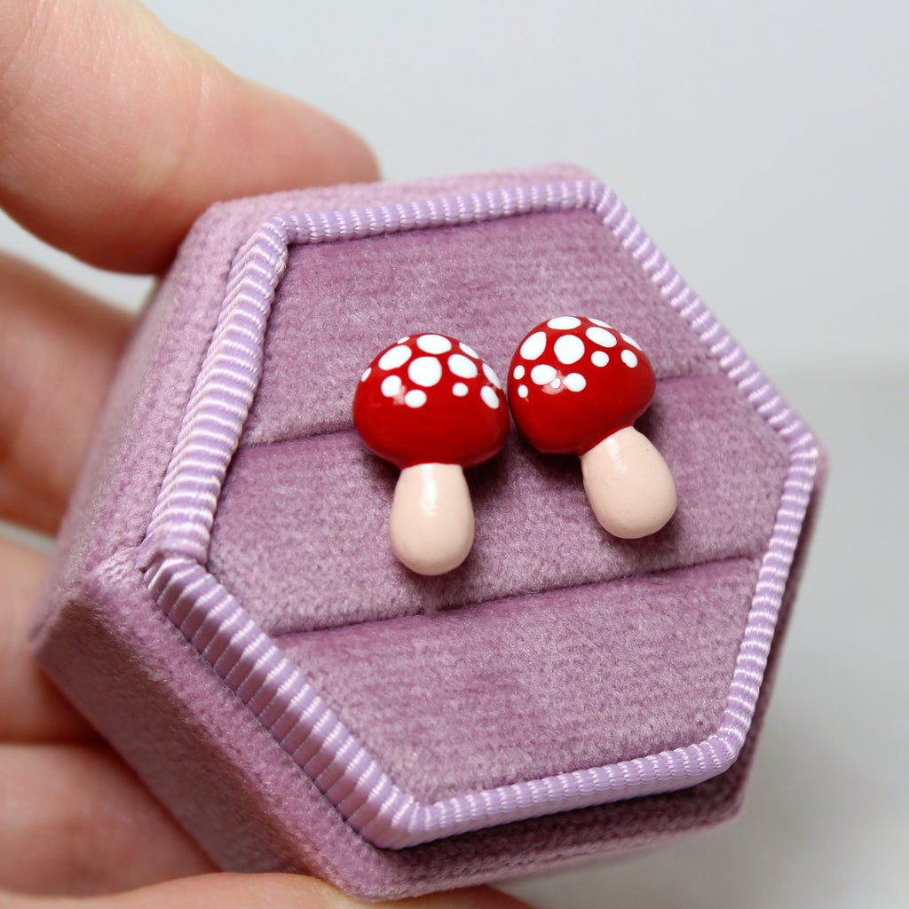 A hand holds a hexagonal purple velvet jewelry box. A pair of red mushroom earrings are nestled in the jewelry box.