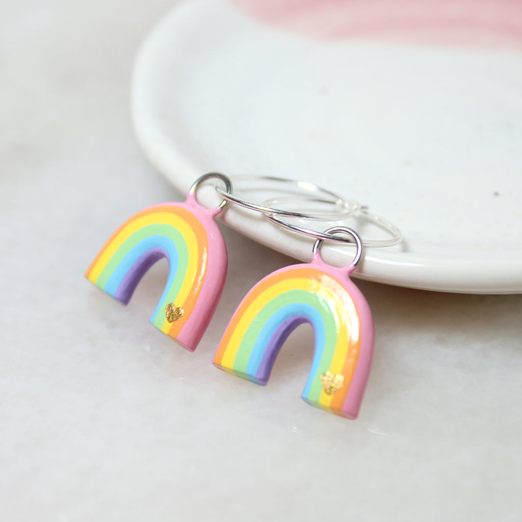 A pair of pastel rainbow charms hang from sterling silver hoop earrings, and rest against a white jewelry dish