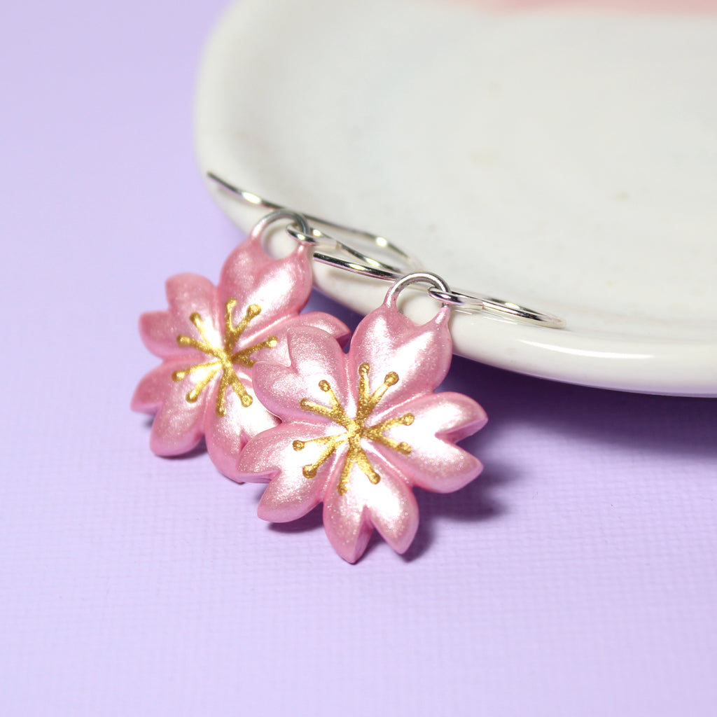 A pair of sparkly cherry blossom charms hang from sterling silver earring hooks. The earrings rest against a white jewelry dish.