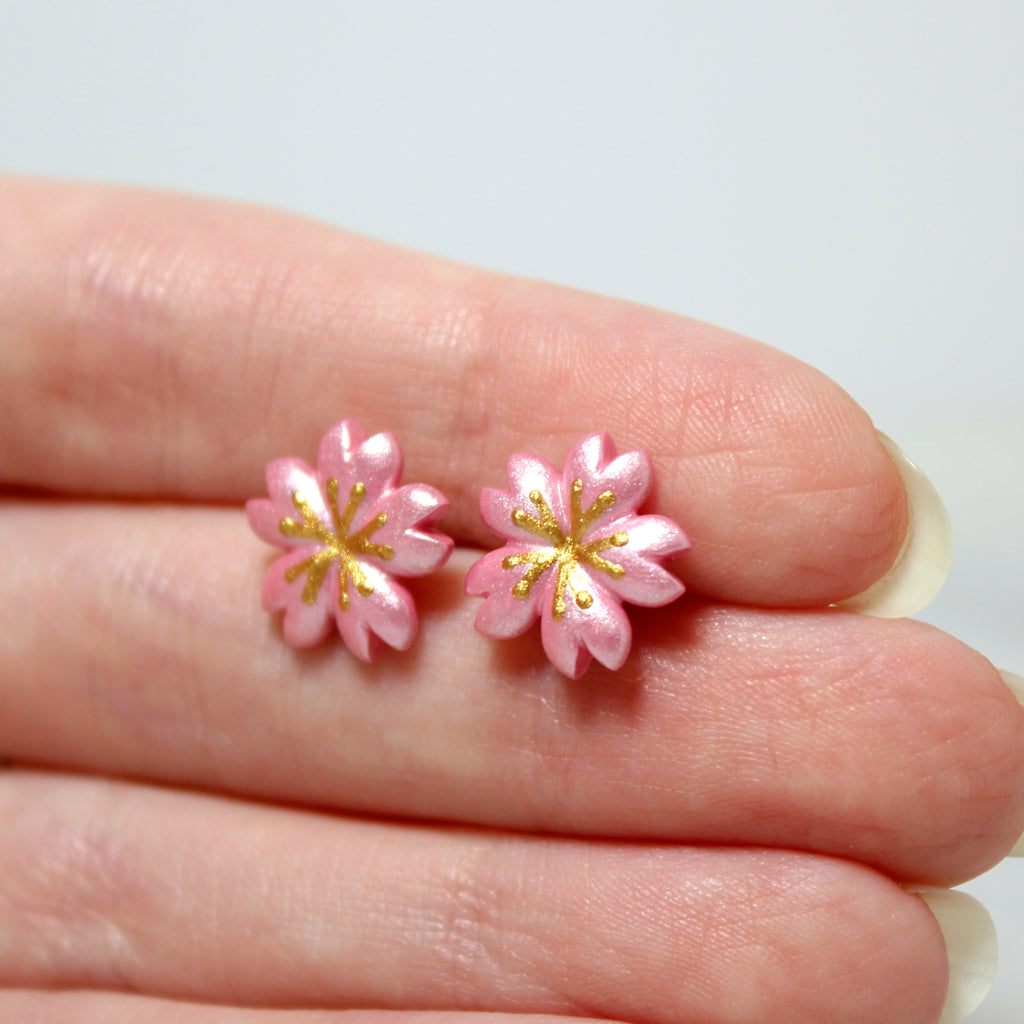 A hand holding a pair of cherry blossom earrings. Each earring is about 1 centimeter wide.