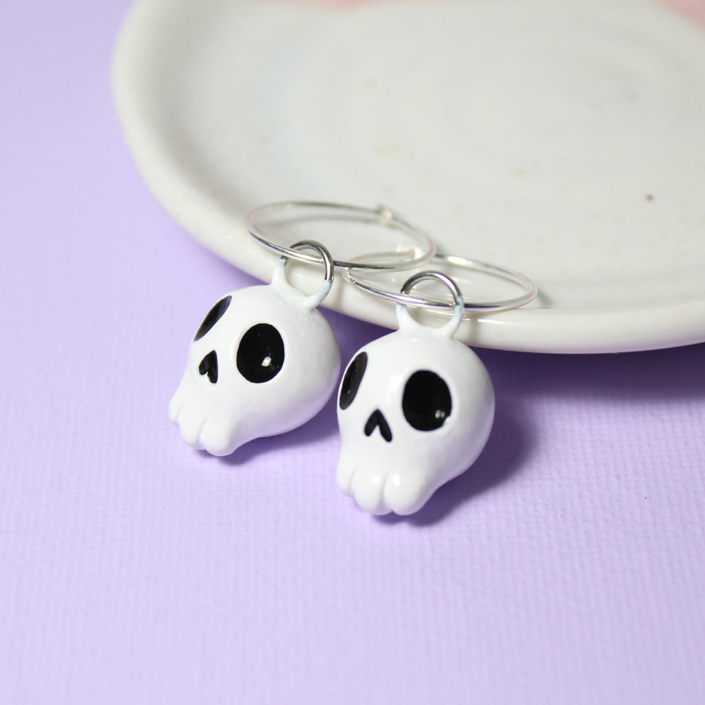 A pair of skull charms hang from sterling silver hoop earrings. The earrings rest against a white jewelry dish on a purple background