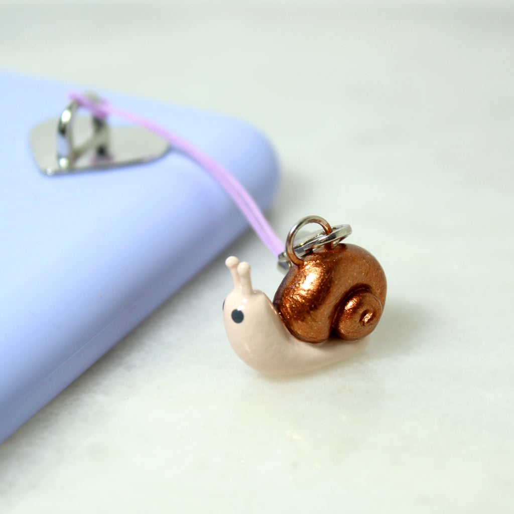 A miniature snail charm with a sparkly bronze shell hangs off of a purple phone case