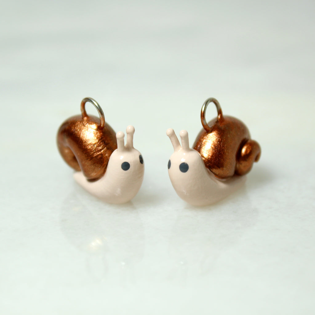 Two miniature snail charms with swirled bronze shells sit next to each other on a marble surface