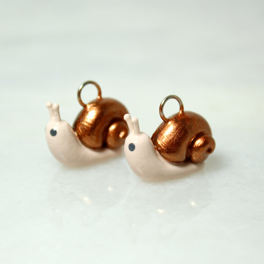 Two miniature snail charms with swirled bronze shells sit next to each other on a marble surface